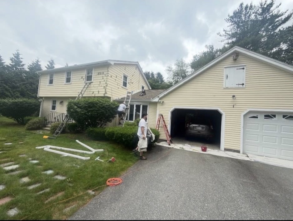 Two-story home with faded yellow siding scraped and primed in preparation for painting, ladders and workers at the wall